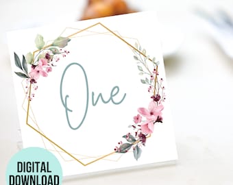 May include: A white table number card with a gold geometric frame and pink and green floral accents. The number "One" is written in blue script.