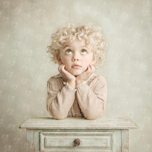 May include: A young child with curly blonde hair gazes upward, resting their chin on clasped hands. They wear a light brown sweater with a lace collar. The child leans on a small, distressed, light blue table with a drawer and a round knob.