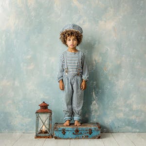 Puede incluir: Un niño con un atuendo de marinero a rayas, que incluye gorro, camisa y pantalón con tirantes. El niño está de pie sobre un baúl azul vintage. Una linterna roja y marrón con una vela encendida está cerca.