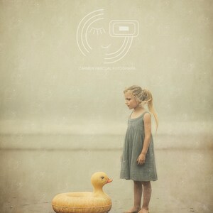 Puede incluir: Una joven se encuentra en una playa junto a un gran pato inflable amarillo. La persona lleva un vestido gris. El pato tiene un cuerpo redondo y una cabeza detallada con un pico naranja. La escena se desarrolla en una playa con una paleta de colores suaves y apagados.