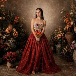 Puede incluir: Un vestido sin tirantes con un corpiño adornado con flores de tela multicolores y una falda de satén rojo oscuro hasta el suelo. La prenda se combina con un pequeño ramo de flores naranjas que se sostiene frente a la cintura.