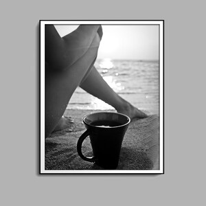 Könnte beinhalten: Schwarzweißfoto einer Person, die auf einem Strand mit einer Tasse Kaffee sitzt. Die Beine der Person sind übereinandergeschlagen und ihre Füße befinden sich im Sand. Das Meer ist im Hintergrund und die Sonne geht unter.
