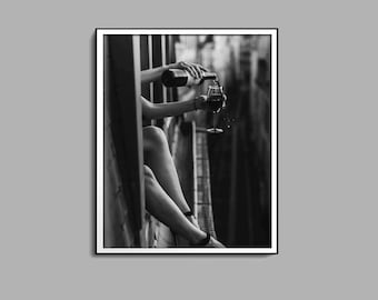 Peut inclure: Une photo en noir et blanc d'une personne versant du vin rouge d'une bouteille dans un verre. La personne est assise sur un rebord et le verre est tenu dans sa main.