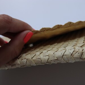 May include: A close-up of a woven straw clutch bag. The bag is folded in half, revealing the woven texture. The bag is a light beige colour.