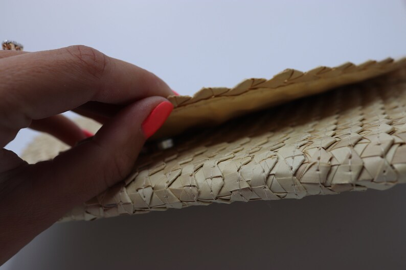 May include: A close-up of a woven straw clutch bag. The bag is folded in half, revealing the woven texture. The bag is a light beige colour.