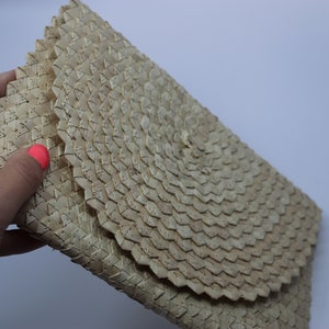 May include: A woven clutch bag made from natural materials, featuring a circular pattern in the centre. The bag is beige and has a slightly textured surface.