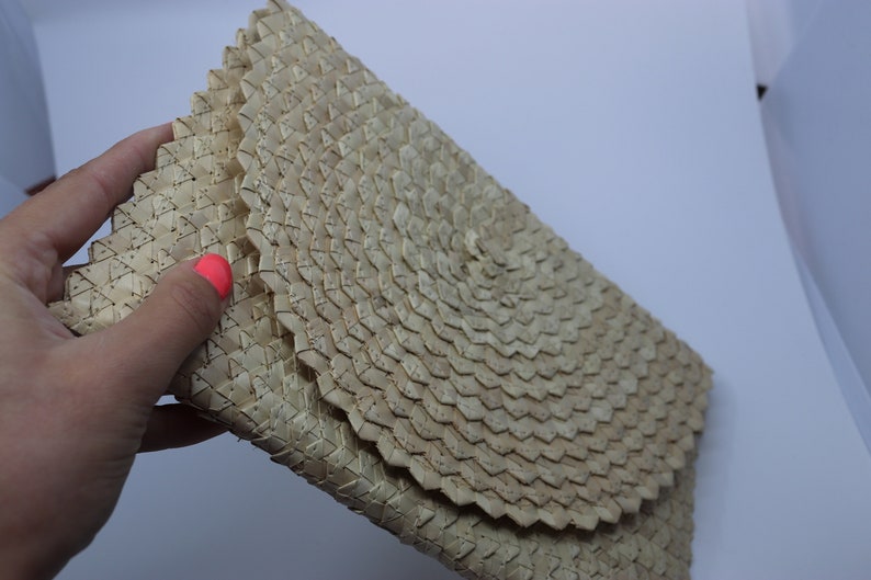 May include: A woven clutch bag made from natural materials, featuring a circular pattern in the centre. The bag is beige and has a slightly textured surface.