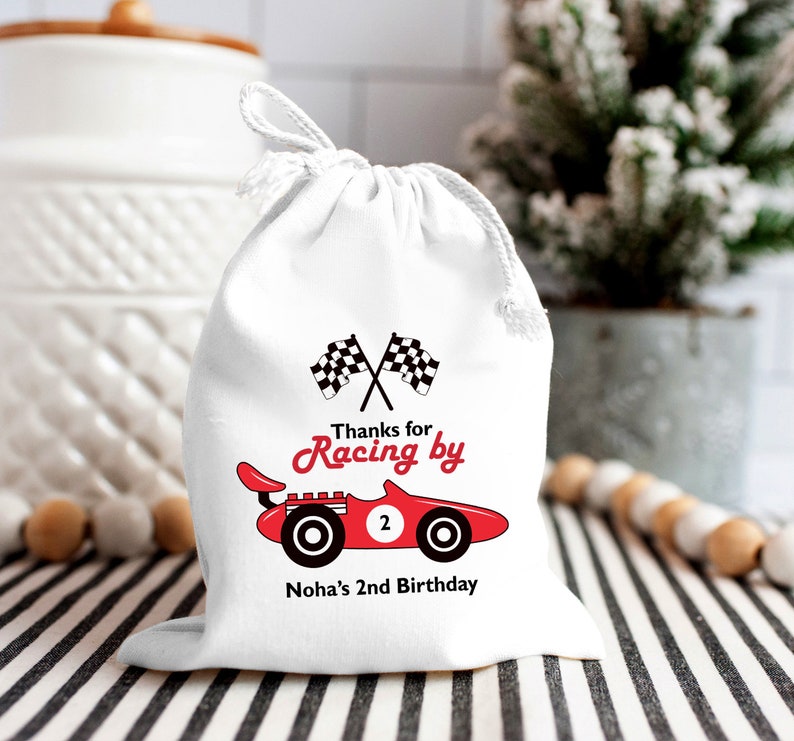 May include: White favor bag with a red race car and checkered flags. The text reads "Thanks for Racing by Noha's 2nd Birthday".