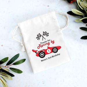 May include: White cotton favor bag with a red race car and checkered flags. The text reads "Thanks for Racing by Noha's 2nd Birthday".