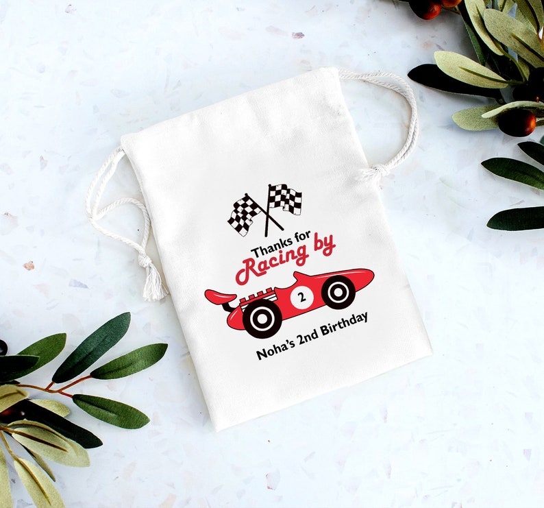 May include: White cotton favor bag with a red race car and checkered flags. The text reads "Thanks for Racing by Noha's 2nd Birthday".