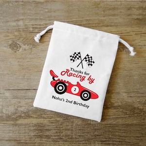 May include: White cotton drawstring bag with a red race car and checkered flags. The text on the bag reads "Thanks for Racing by Noha's 2nd Birthday."