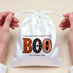 May include: White satin drawstring bag with the word "BOO" in orange and black Halloween-themed lettering. The letter "B" has orange polka dots, the "O" has a spider, and the last "O" has a pumpkin. Text reads "A Spooky Treat Just For You!" and "Your Friend, Alex."