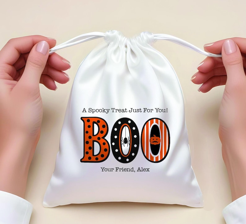 May include: White satin drawstring bag with the word "BOO" in orange and black Halloween-themed lettering. The letter "B" has orange polka dots, the "O" has a spider, and the last "O" has a pumpkin. Text reads "A Spooky Treat Just For You!" and "Your Friend, Alex."