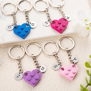 Könnte beinhalten: Schlüsselanhänger mit herzförmigen Anhängern in Blau, Pink, Lila und Hellrosa. Jeder Schlüsselanhänger hat einen silbernen Ring, eine Kette und einen kleinen runden Anhänger mit einem Buchstaben. Die Anhänger sehen aus wie ineinandergreifende Spielzeugbausteine.