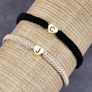 May include: Two friendship bracelets with heart-shaped charms. One bracelet is black with a gold charm with the letter "C" and the other is beige with a gold charm with the letter "J".