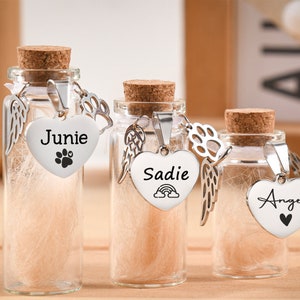 Puede incluir: Tres pequeños frascos de vidrio con tapones de corcho, cada uno con un material de color claro. Cada frasco tiene un dije plateado en forma de corazón con un nombre y una huella de pata o un símbolo de corazón. Un dije dice "Junie", otro "Sadie" y el último "Angel".