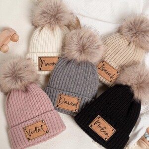 Könnte beinhalten: Eine Auswahl personalisierter Strickmützen in verschiedenen Farben wie Rosa, Grau, Schwarz, Creme und Beige. Jede Mütze hat einen Kunstfell-Bommel und ein Kunstleder-Namensschild. Ein Holzspielzeugauto und eine Rassel sind ebenfalls zu sehen.