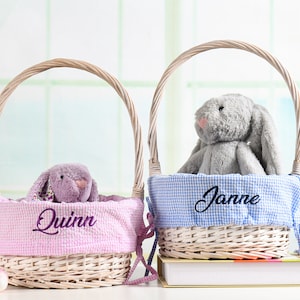 Könnte beinhalten: Zwei weiße Weidenkörbe mit rosa und blauen Gingham-Futter. Die Körbe sind mit Plüsch-Hasenspielzeug gefüllt. Der rosa Korb hat den Namen "Quinn" bestickt, und der blaue Korb hat den Namen "Janne" bestickt.