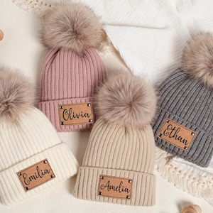 Könnte beinhalten: Vier gestrickte Mützen mit Kunstfell-Bommeln. Die Hüte sind in Creme, Rosa, Beige und Grau gehalten, jede mit einem Namensschild aus Kunstleder. Die Namen auf den Etiketten lauten "Emilia", "Olivia", "Amelia" und "Ethan".