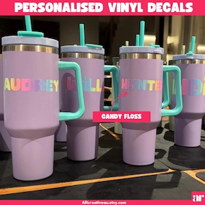 May include: A collection of lavender insulated tumblers with teal handles and straws. Each tumbler features a name in rainbow-coloured lettering. The image includes the text "PERSONALIZED VINYL DECALS" and the text "CANDY FLOSS".