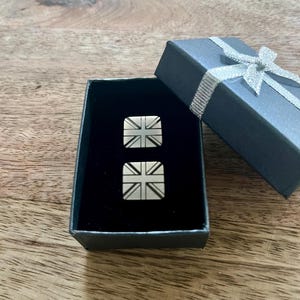 May include: A pair of silver cufflinks in a black box with a silver ribbon. The cufflinks feature a design of the Union Jack flag.