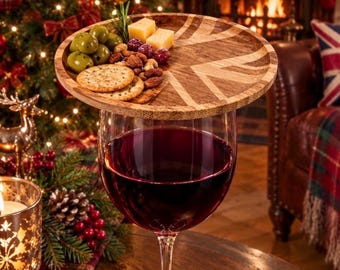 May include: A round wooden snack board featuring a Union Jack pattern, designed to sit atop a wine glass. The board holds a selection of green olives, crackers, nuts, and cheese cubes, while the glass below is filled with red wine.