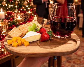 May include: A wooden serving board holding a glass of red wine, a wedge of brie cheese, square crackers, green grapes, cubes of cheddar cheese, and fresh strawberries. The board features a circular indentation for the wine glass base.