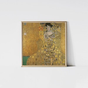 May include: A framed print of a Gustav Klimt painting. The artwork features a woman in a long, ornate gold dress with intricate patterns. The background is also gold, with geometric shapes and textures. The frame is gold.