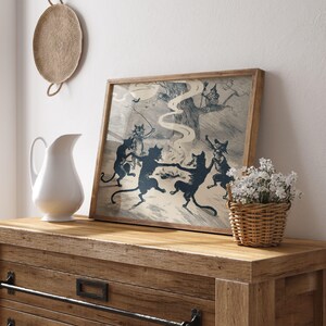 May include: A framed print featuring a vintage illustration of dancing black cats in a circle, with a witch in a tree. The artwork has a light background and is set in a wooden frame. A white pitcher and a basket of flowers are on a wooden dresser.