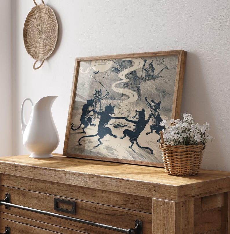 May include: A framed print featuring a vintage illustration of dancing black cats in a circle, with a witch in a tree. The artwork has a light background and is set in a wooden frame. A white pitcher and a basket of flowers are on a wooden dresser.