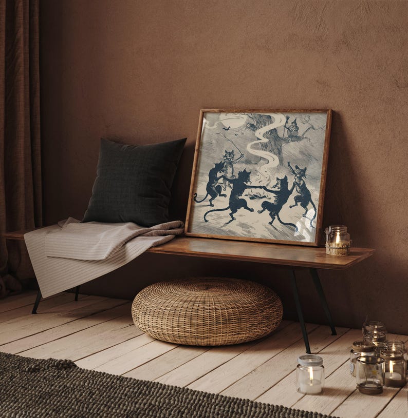 May include: A framed print depicts dancing black cats around a bonfire, with a full moon in the background. The print is displayed on a wooden bench with a dark gray pillow and a striped blanket. A woven ottoman and candles complete the scene.