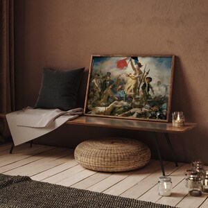 May include: A framed print of a classic painting depicting a historical scene with figures and a flag. The artwork is displayed on a wooden bench with a dark gray pillow and a light gray blanket. A woven ottoman and candles are in the foreground.