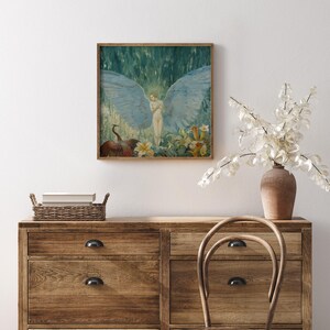 May include: A framed artwork depicts a nude figure with large blue wings, surrounded by flowers and a bird. The painting has a vintage aesthetic with a wooden frame. The artwork is displayed above a wooden dresser with a woven basket and a vase of white flowers.