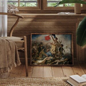 May include: A framed print of a classic painting, featuring a woman holding a flag amidst a battle scene. The artwork is displayed in a room with wooden floors, a woven chair, and a neutral-toned throw. A book and a plant are also visible.