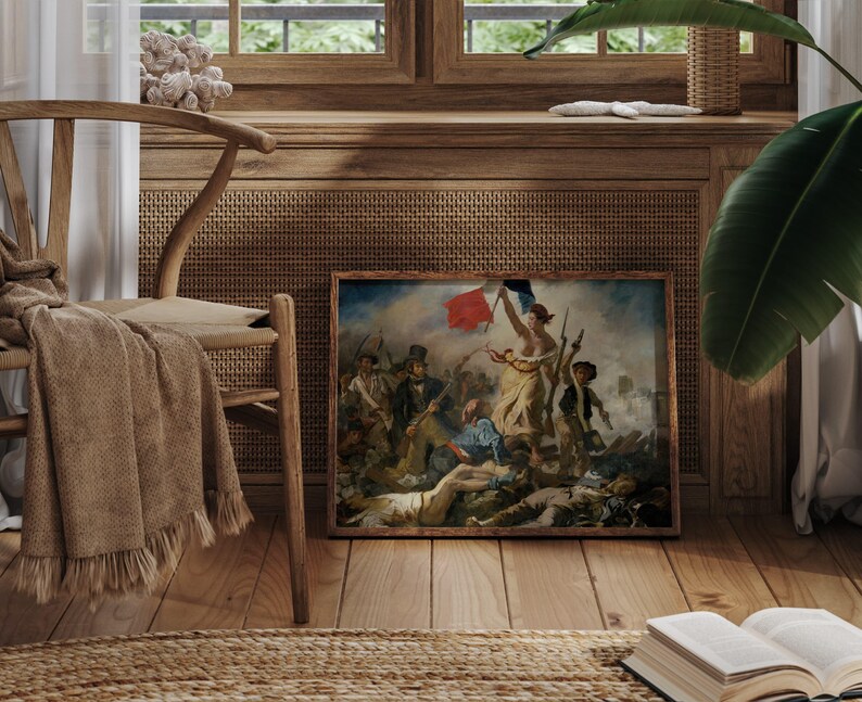 May include: A framed print of a classic painting, featuring a woman holding a flag amidst a battle scene. The artwork is displayed in a room with wooden floors, a woven chair, and a neutral-toned throw. A book and a plant are also visible.