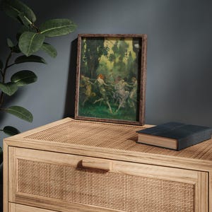 May include: A framed artwork depicting figures dancing in a forest setting. The painting features a palette of greens, yellows, and blues, with a dark brown wooden frame. The artwork rests on a light brown woven surface, with a closed book nearby.