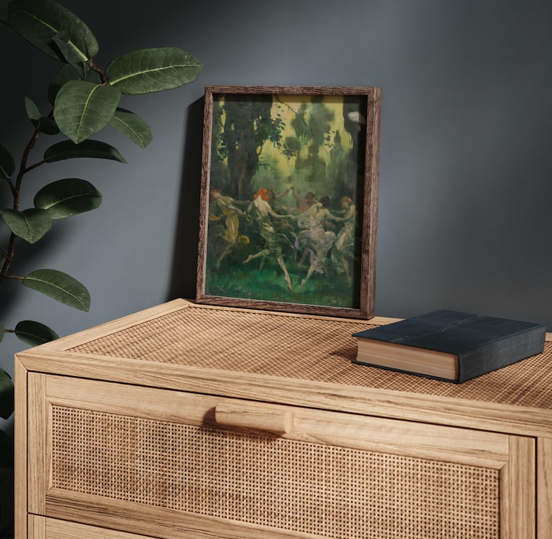 May include: A framed artwork depicting figures dancing in a forest setting. The painting features a palette of greens, yellows, and blues, with a dark brown wooden frame. The artwork rests on a light brown woven surface, with a closed book nearby.