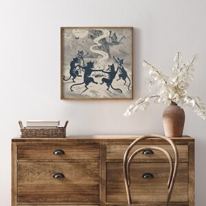 May include: A framed print depicts a whimsical scene of dancing black cats in a forest setting. The artwork, in shades of blue and cream, features a witch on a tree and bats. The print is displayed above a wooden dresser with a vase of white flowers.