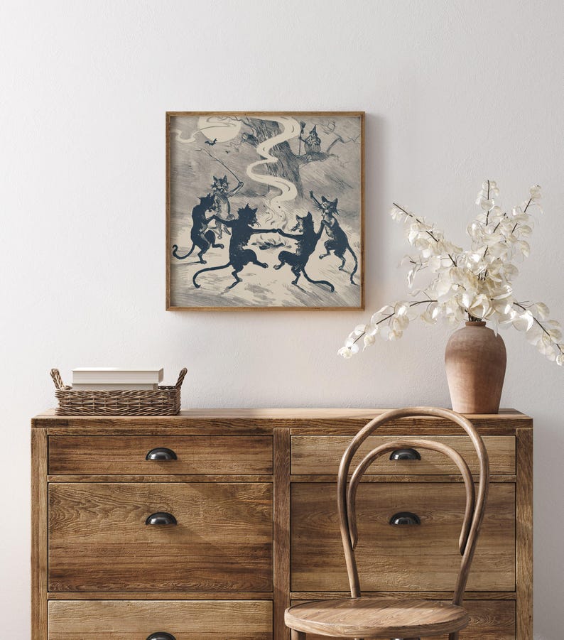 May include: A framed print depicts a whimsical scene of dancing black cats in a forest setting. The artwork, in shades of blue and cream, features a witch on a tree and bats. The print is displayed above a wooden dresser with a vase of white flowers.