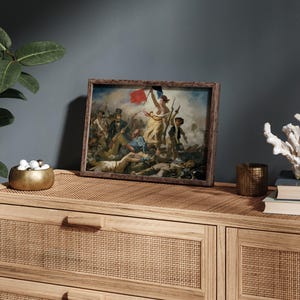 May include: A framed print of a classic painting depicting a historical scene with figures, a flag, and a woman holding a flag. The print is in a brown wooden frame, displayed on a light brown wooden cabinet. Decorative items include a gold bowl, a candle holder, and a white coral.