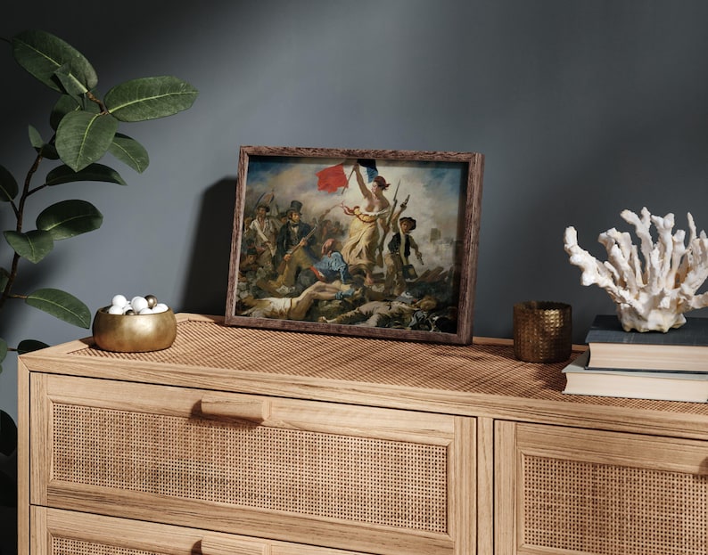 May include: A framed print of a classic painting depicting a historical scene with figures, a flag, and a woman holding a flag. The print is in a brown wooden frame, displayed on a light brown wooden cabinet. Decorative items include a gold bowl, a candle holder, and a white coral.