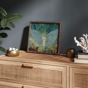 May include: A framed artwork featuring an angel with large blue wings, set against a backdrop of green and white. The artwork is displayed on a light wood cabinet with a woven front, alongside a gold bowl, candle holder, and coral.