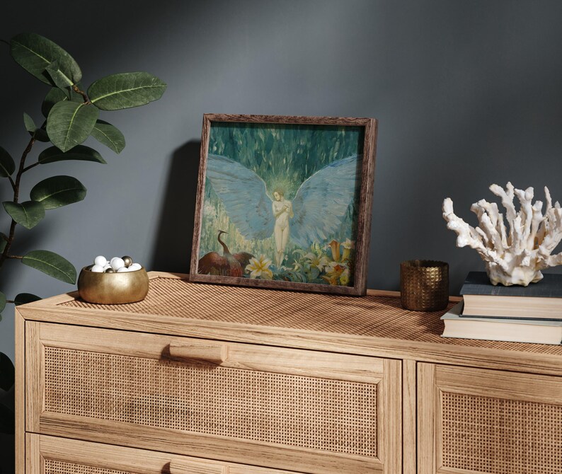 May include: A framed artwork featuring an angel with large blue wings, set against a backdrop of green and white. The artwork is displayed on a light wood cabinet with a woven front, alongside a gold bowl, candle holder, and coral.