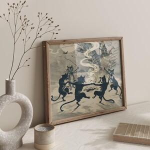 May include: A framed print featuring a vintage illustration of dancing black cats in a spooky scene. The artwork has a light blue and beige color palette, with a wooden frame. The print depicts a Halloween or witch-themed gathering.