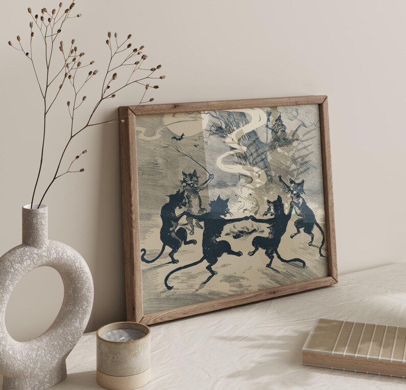 May include: A framed print featuring a vintage illustration of dancing black cats in a spooky scene. The artwork has a light blue and beige color palette, with a wooden frame. The print depicts a Halloween or witch-themed gathering.