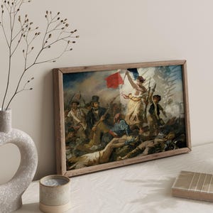 May include: A framed print of a historical painting depicting a battle scene with figures, a red flag, and a woman holding a flag. The artwork is in a wooden frame, leaning against a wall. A vase with branches and a candle are in the foreground.