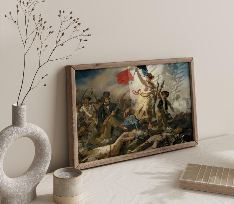 May include: A framed print of a historical painting depicting a battle scene with figures, a red flag, and a woman holding a flag. The artwork is in a wooden frame, leaning against a wall. A vase with branches and a candle are in the foreground.