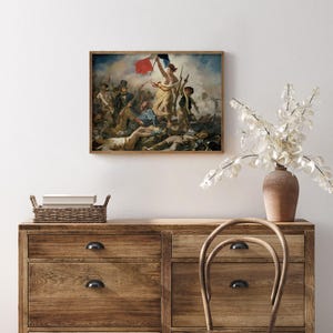 May include: A framed print of a historical painting depicting a battle scene with figures holding flags. The artwork features a woman holding a flag, surrounded by soldiers and fallen figures. The frame is a warm brown color, and the painting is displayed on a white wall.