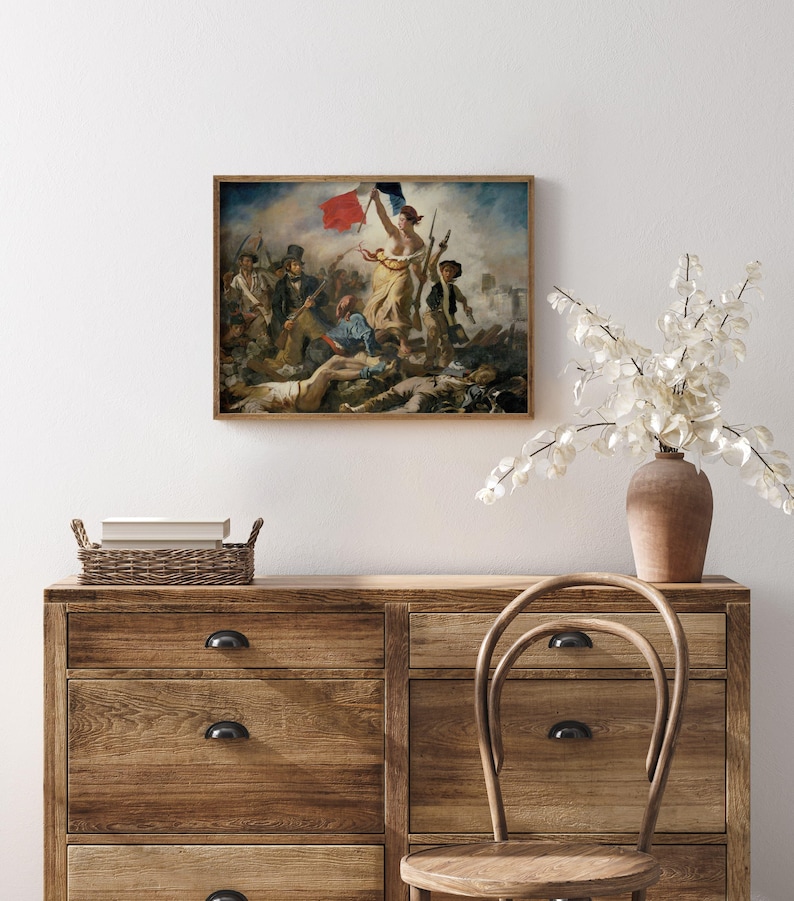 May include: A framed print of a historical painting depicting a battle scene with figures holding flags. The artwork features a woman holding a flag, surrounded by soldiers and fallen figures. The frame is a warm brown color, and the painting is displayed on a white wall.