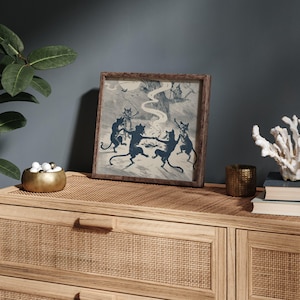 May include: A framed print featuring a vintage illustration of dancing black cats in a forest setting. The artwork is in shades of blue and gray, set in a wooden frame. The print is displayed on a light wood cabinet.